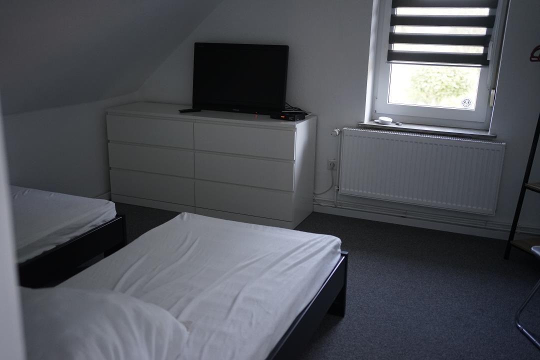 Two beds, dresser with TV, window with blinds, and radiator.