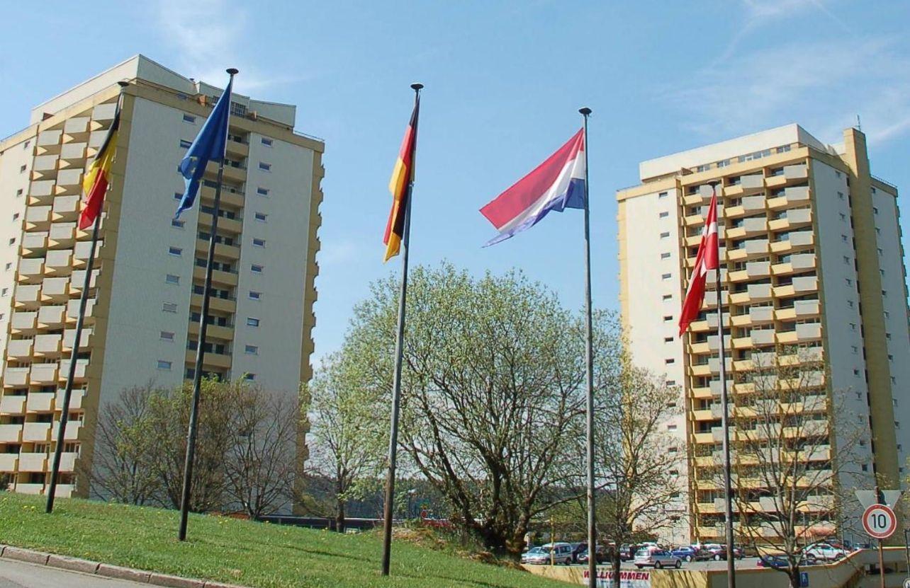 Zwei Hochhäuser mit Flaggen und Bäumen im Vordergrund. Parkplatz mit Autos.