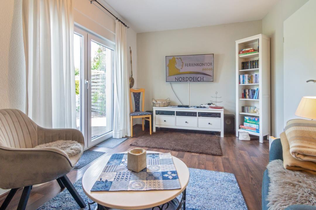 Wohnzimmer mit Sofa, Sessel, Tisch und Fernseher. Fenster mit Blick auf Terrasse.