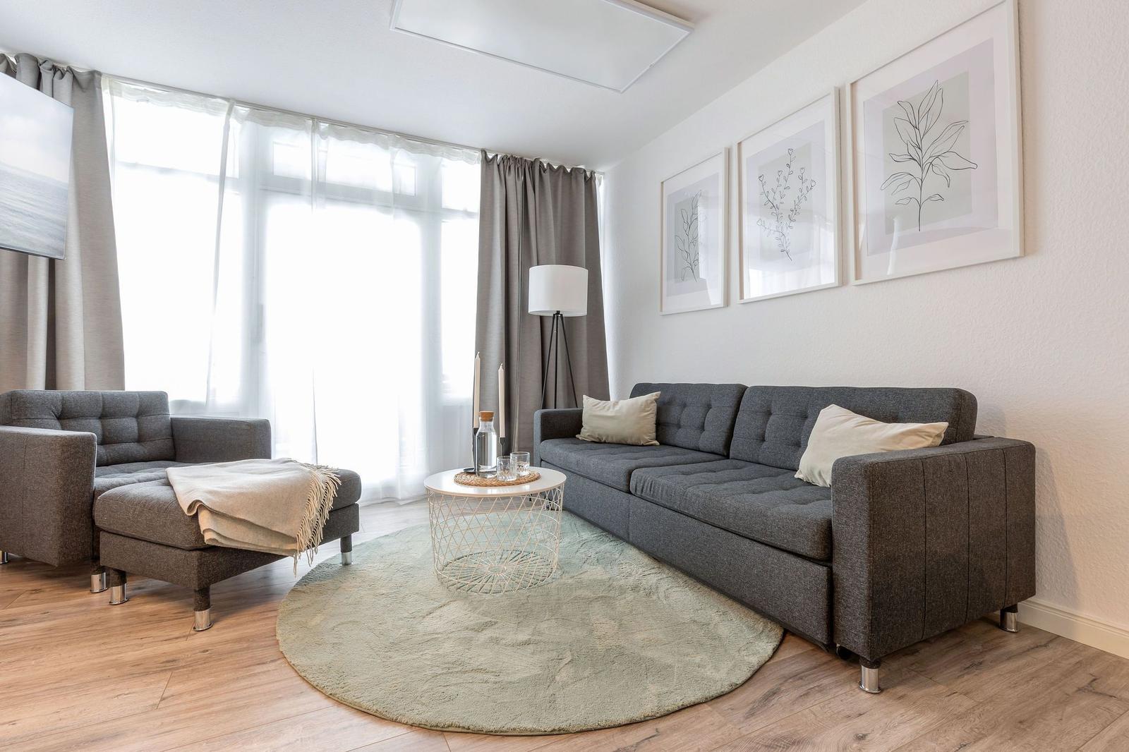 Wohnzimmer mit grauem Sofa, runden Teppich und Fenstern mit Vorhängen.