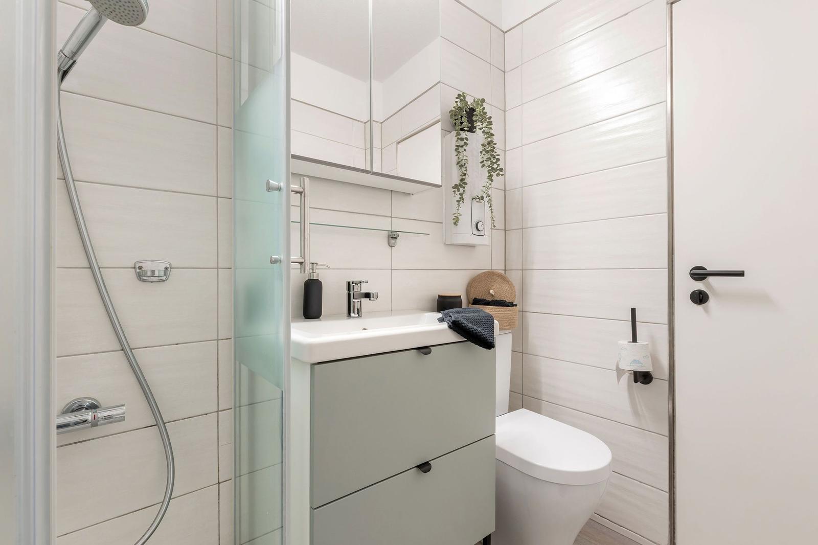 Badezimmer mit Dusche, Waschbecken und Toilette. Weiße Fliesen und graue Schrank.