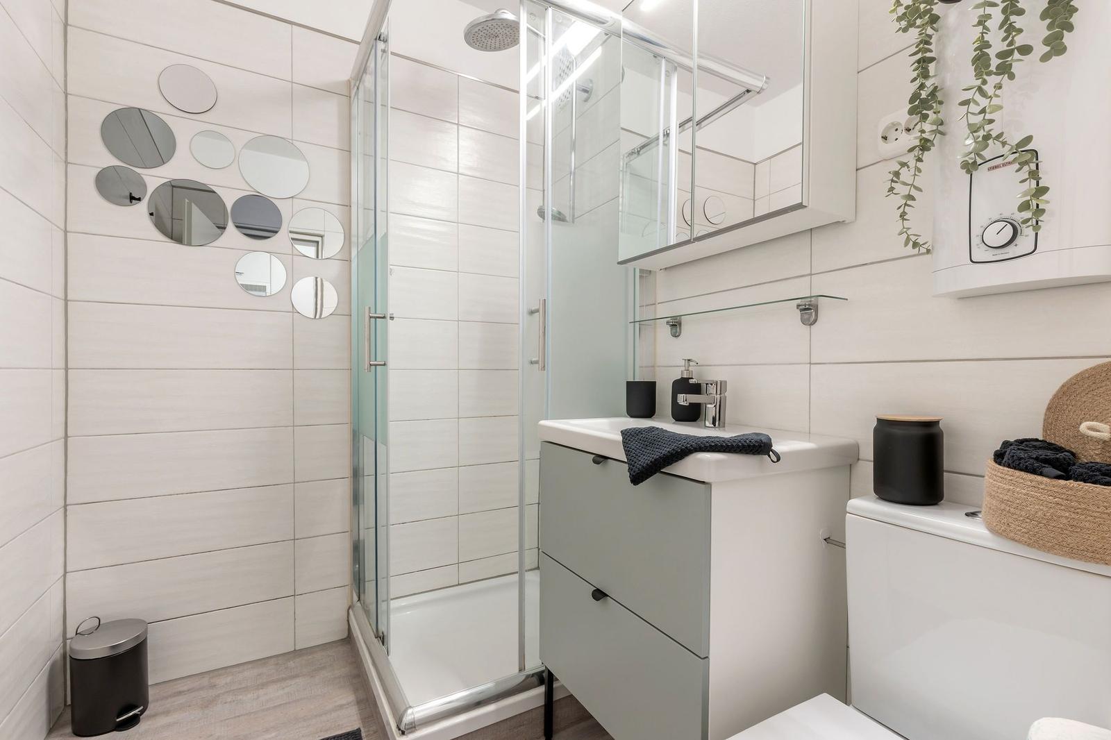 Badezimmer mit Dusche, Waschbecken und Toilette. Wand mit runden Spiegeln und Pflanze.