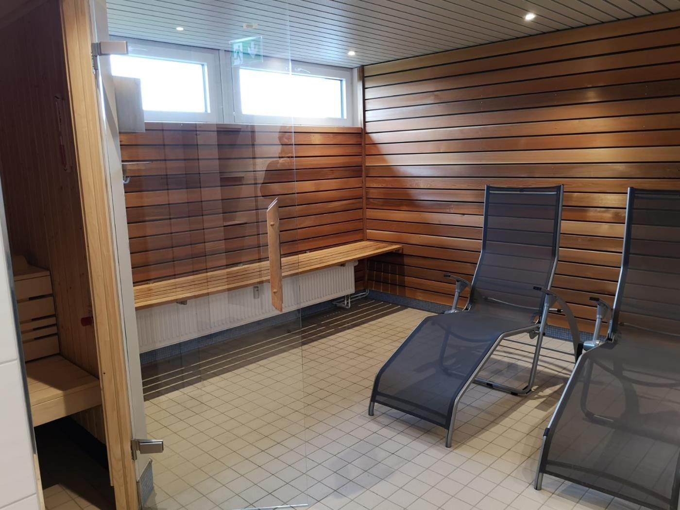 Sauna mit Holzwand, Liegen und Fenster. Tür führt in einen separaten Raum.