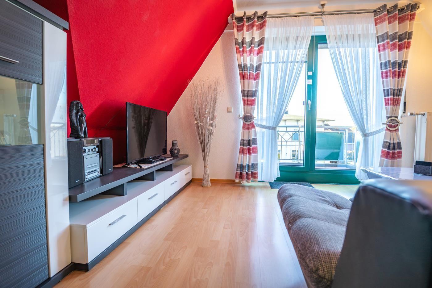 Wohnzimmer mit TV, Sofa und Balkon. Rote Wand und Holzboden.