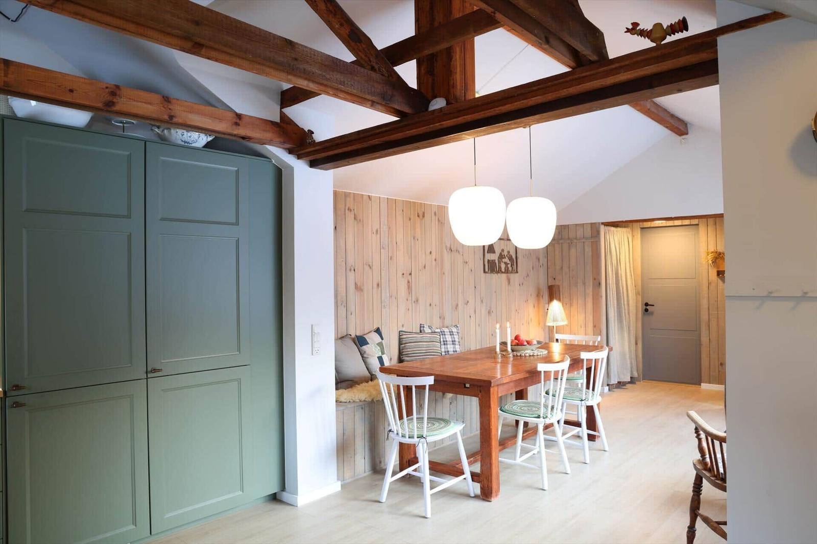 Dining area with wooden table, white chairs, and wood-paneled wall. Exposed beams and green cabinets.