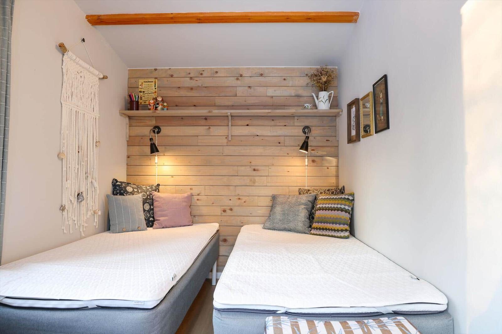 Two beds with pillows are against a wooden wall with shelf and lamps.