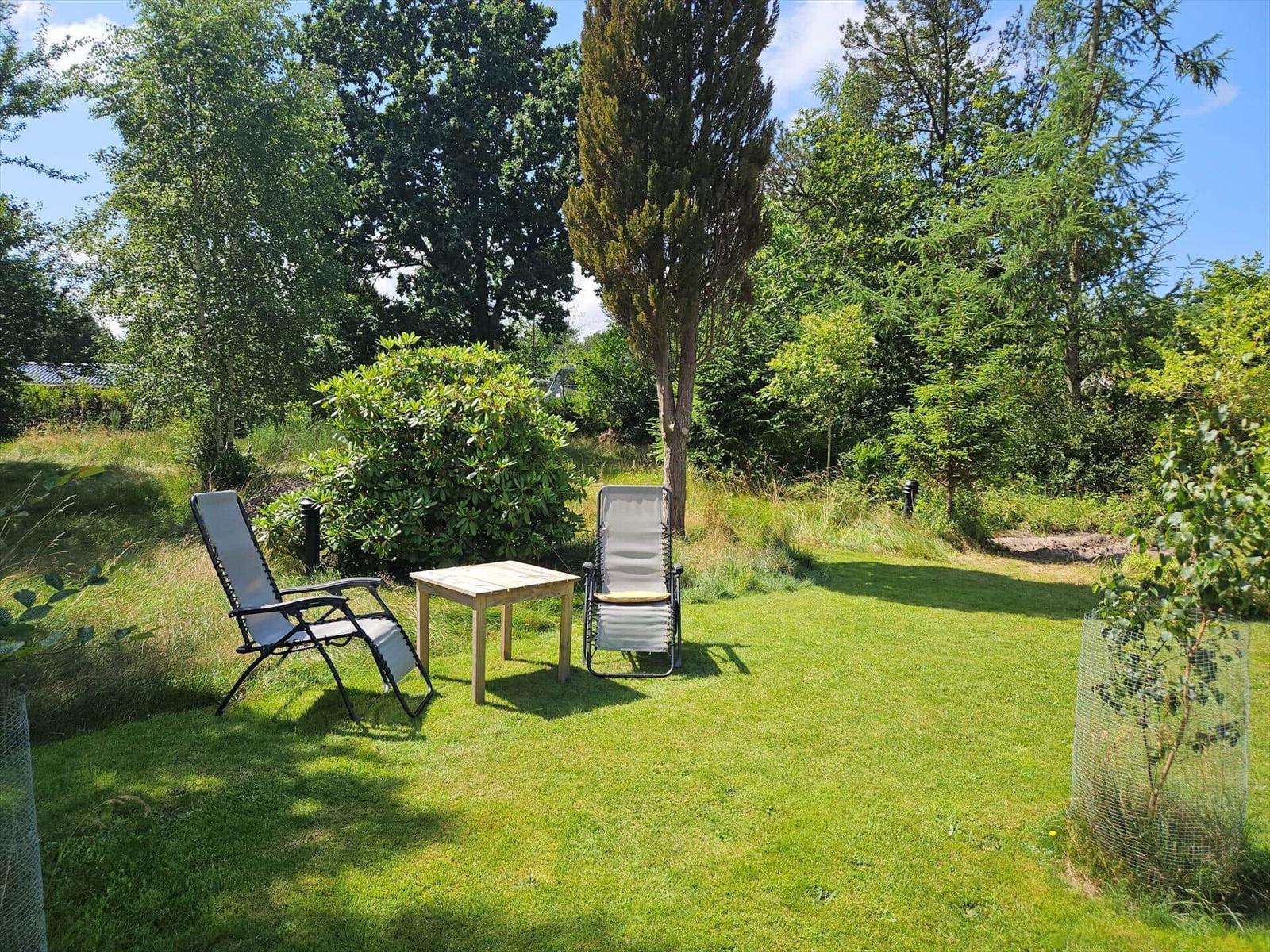 Garden with two recliners and table. Lawn area, trees, and shrubs surround the space.