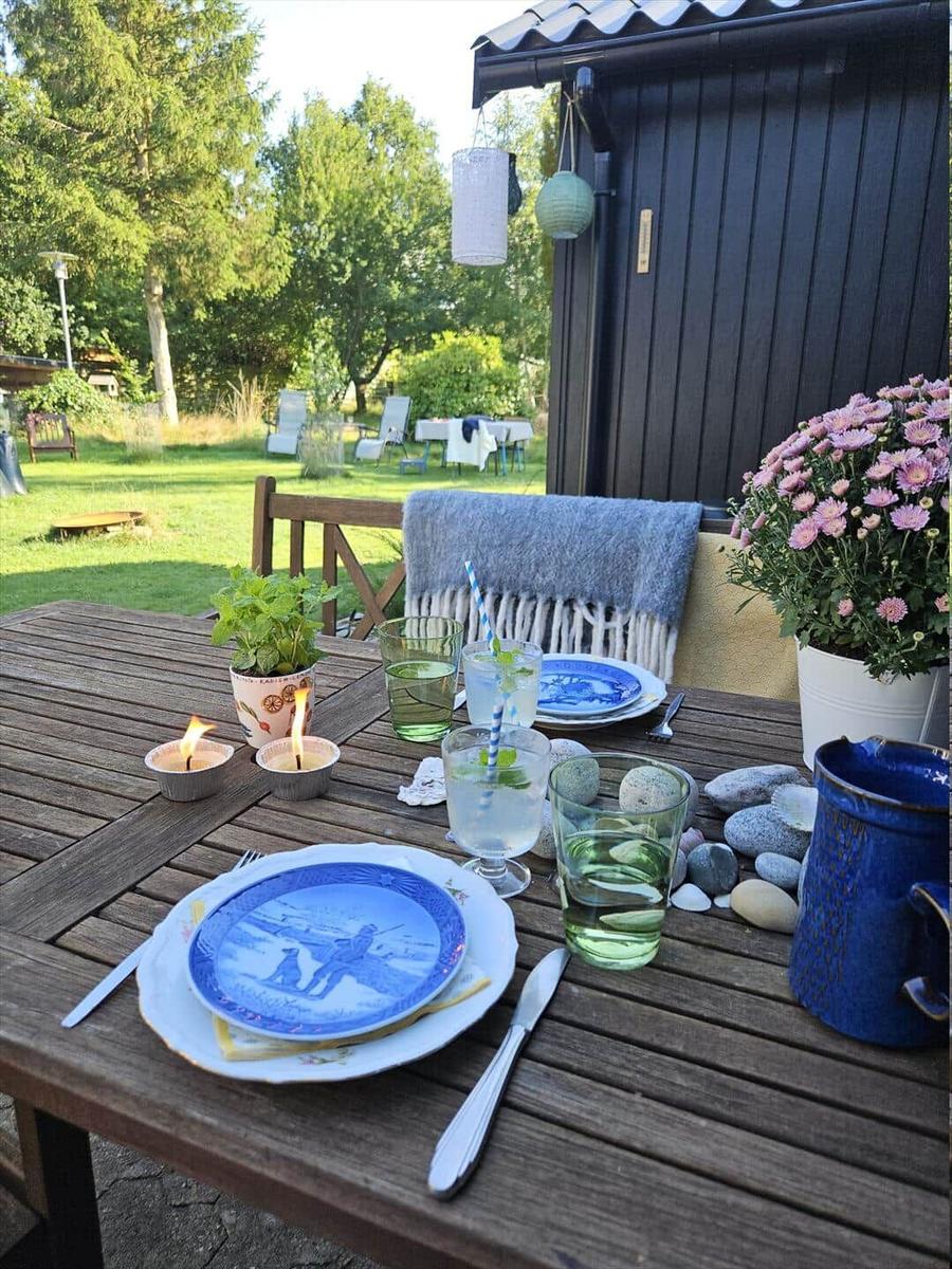Garden table with table setting, flowers, and stones. Background with garden furniture and trees.