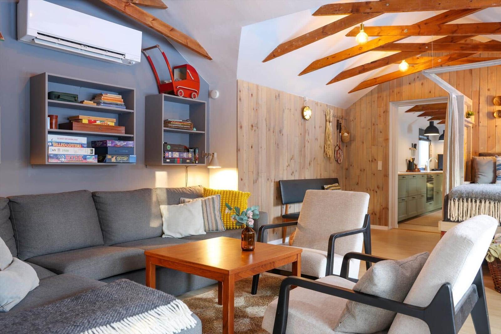 Living room with sofa, chairs, and wooden table. Wooden walls and ceiling beams. View into kitchen and bedroom.