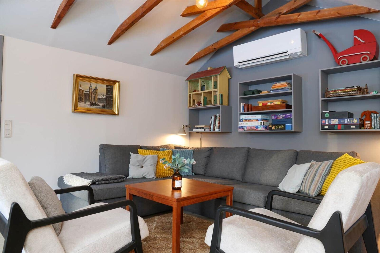 A living room with a gray sofa, two chairs, and a wooden table. Shelves on the wall hold books and games.