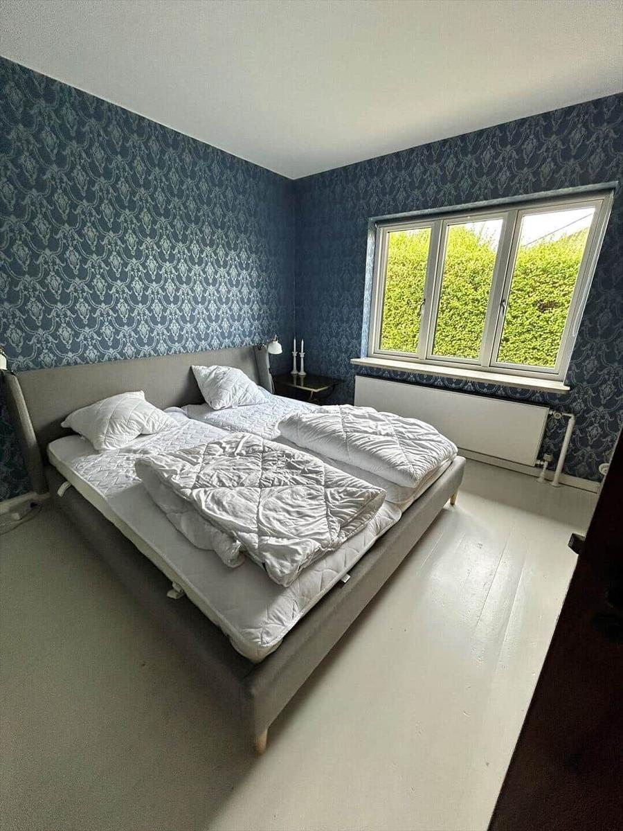 Bedroom with double bed, patterned blue wallpaper, and window with view of green hedge.