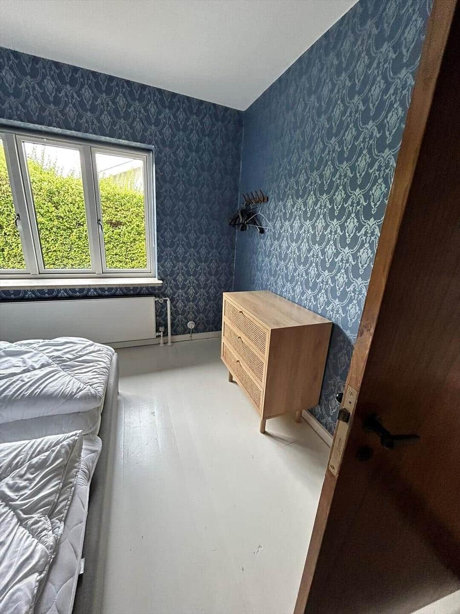 Bedroom with blue patterned wallpaper, bed, dresser, and window overlooking greenery.