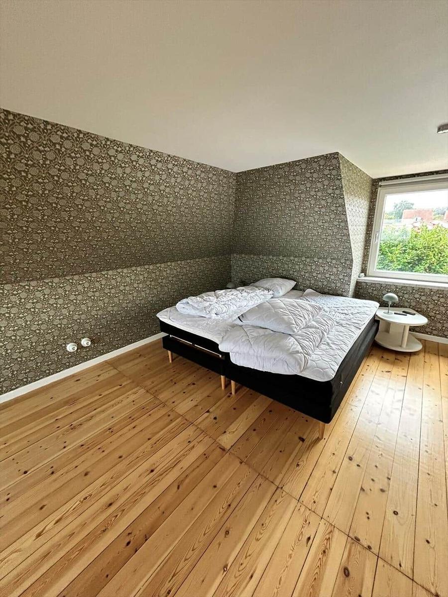 Bedroom with wooden floor, patterned wall covering, and window overlooking greenery.