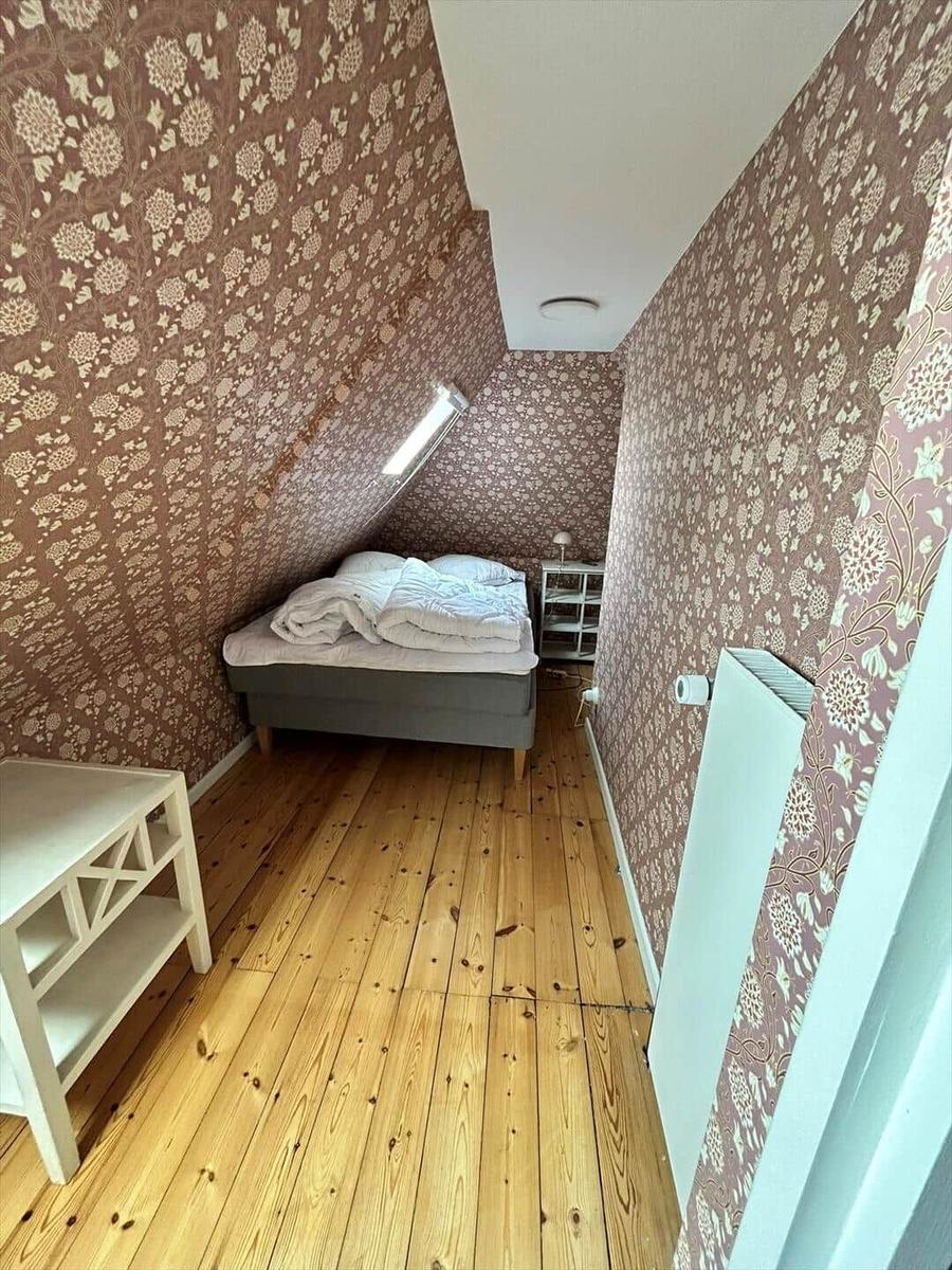Bedroom with wooden floor, patterned wallpaper, and sloped ceiling. Bed, nightstand, and radiator.