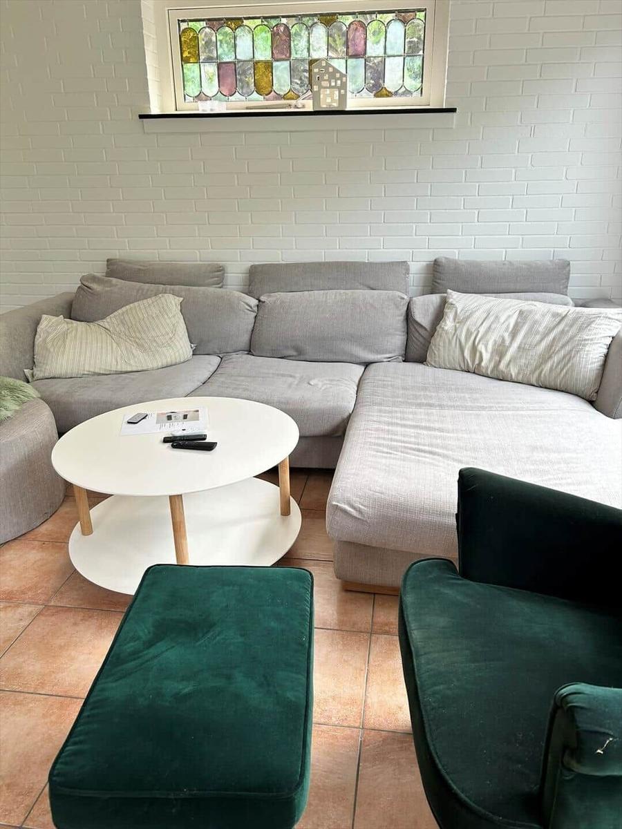 Living room with gray sofa, green armchair, round table. White brick wall and stained glass window.