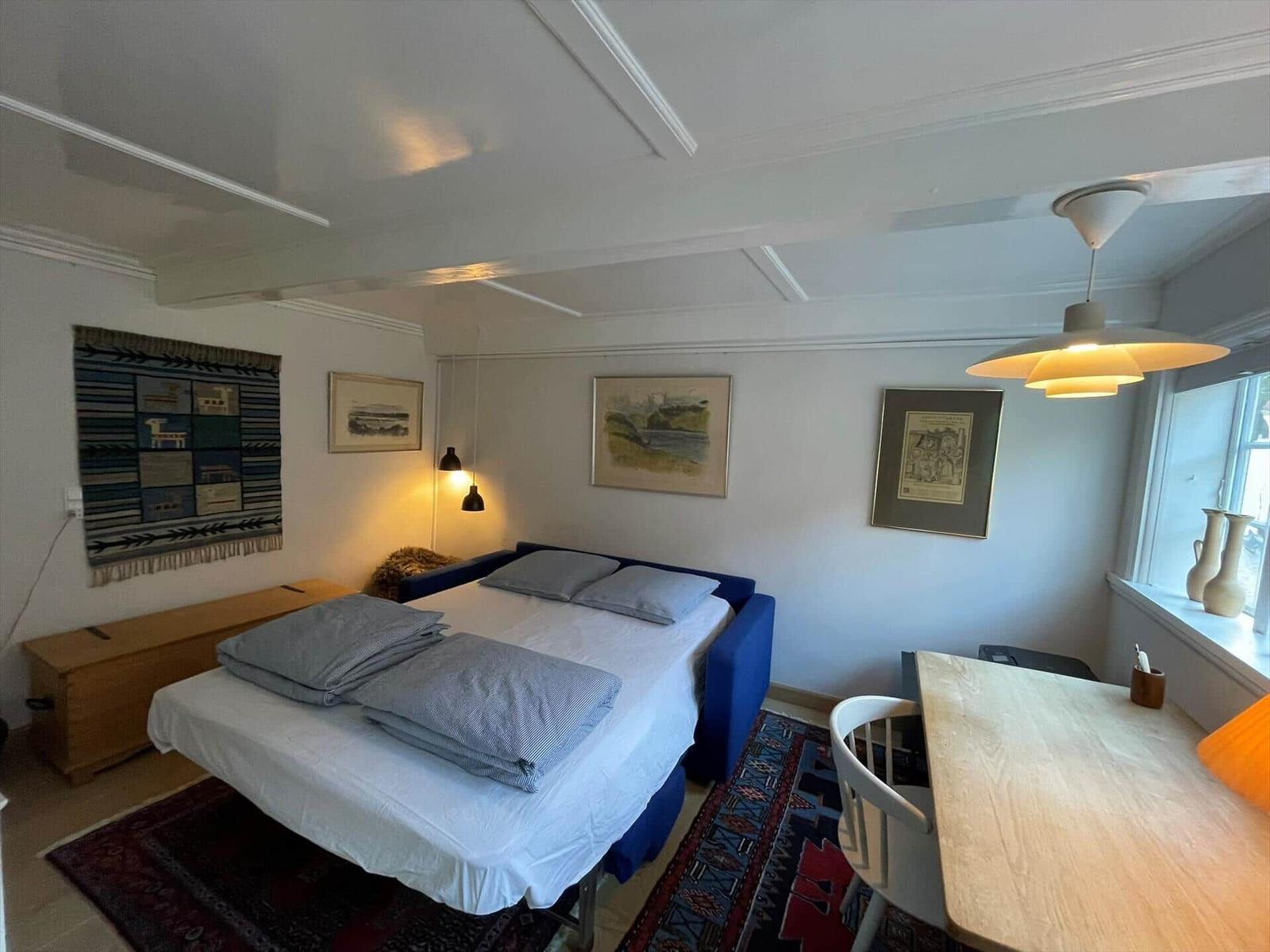 A bedroom with bed, table and window. Wall art and ceiling light are visible.