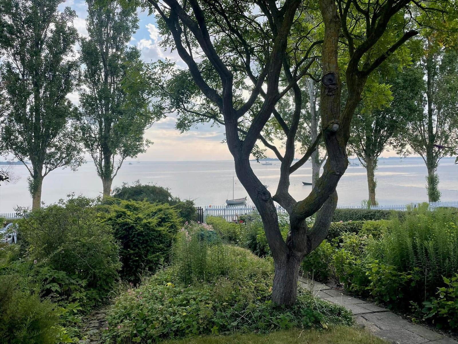 Garden path with trees and view of calm water with boats.