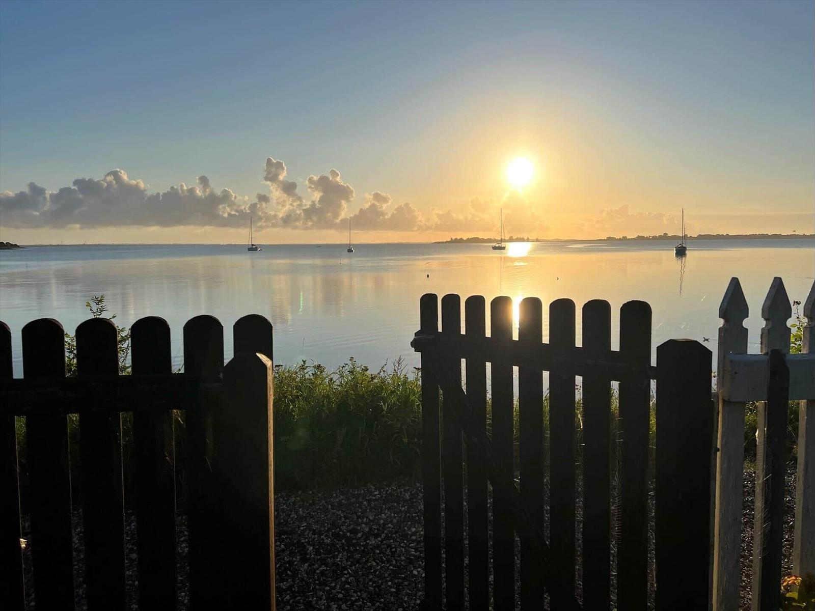 Sunset over calm water with sailboats and a fence in the foreground.