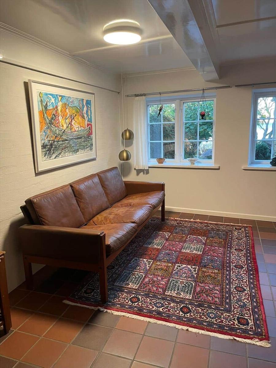 A living room with a brown leather sofa, a colorful rug, and two windows with a view of greenery.