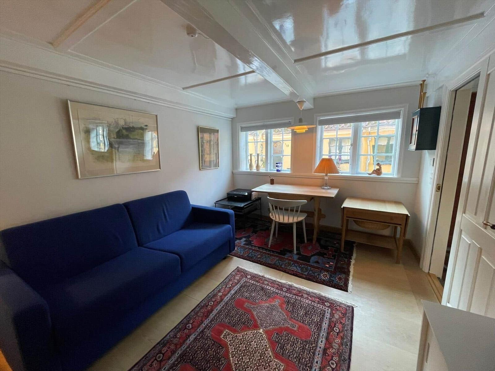 A room with blue sofa, table, chair and two rugs. Pictures hang on the wall.