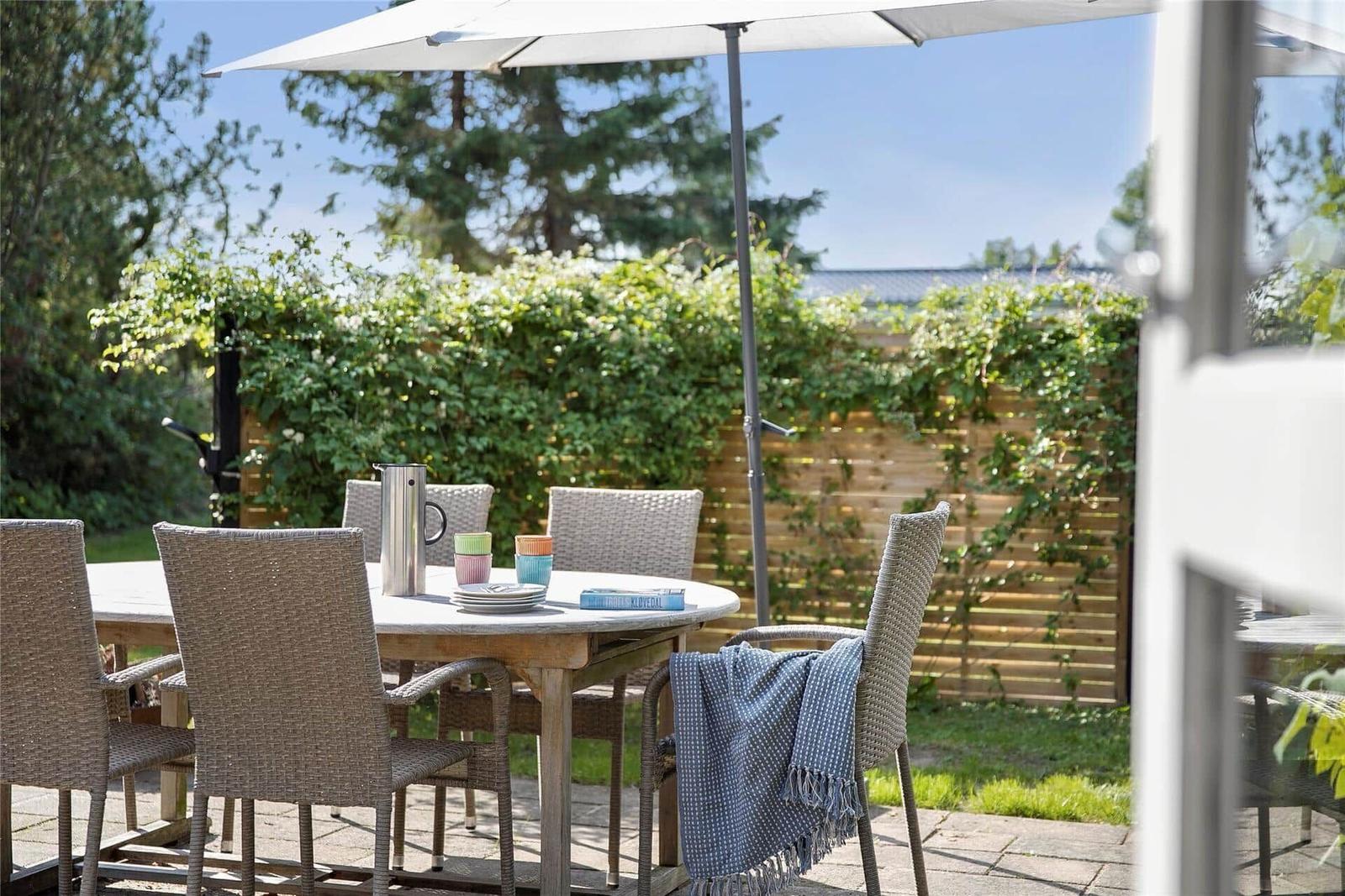 Terrasse mit Tisch, Stühlen und Sonnenschirm. Hintergrund: grüne Hecke und Holzzaun.