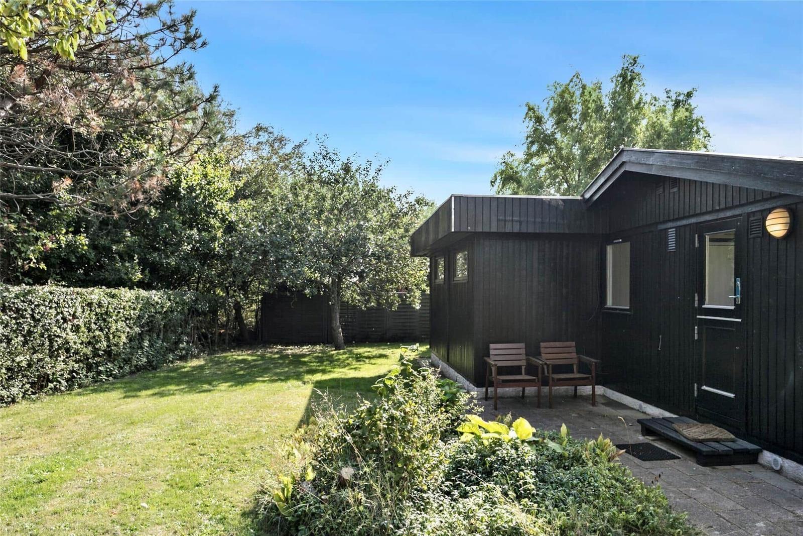 Ein dunkles Holzhaus mit Garten, zwei Stühlen und Bäumen im Hintergrund.