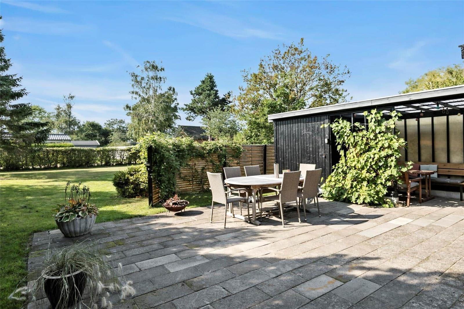 Hinterhof mit Tisch, Stühlen, Grill und Pflanzen. Holzhaus und Rasen im Hintergrund.