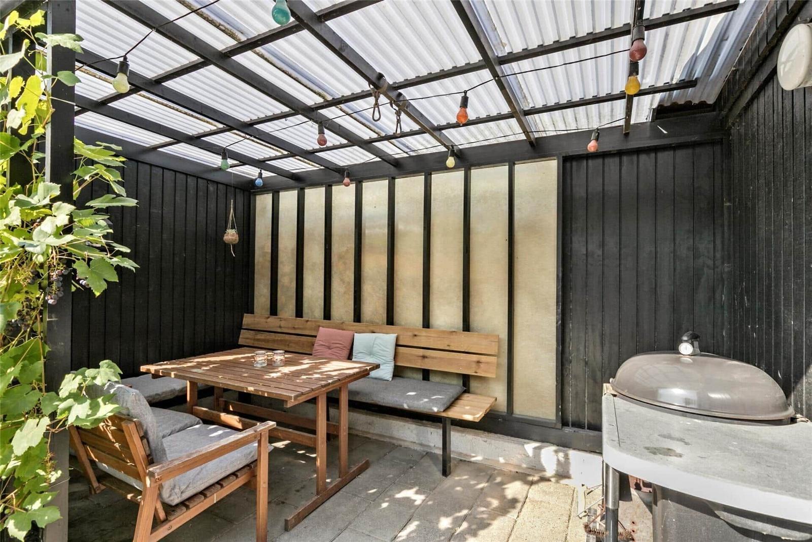 Gartenbereich mit Holztisch, Bank, Grill und Girlanden unter Dach.