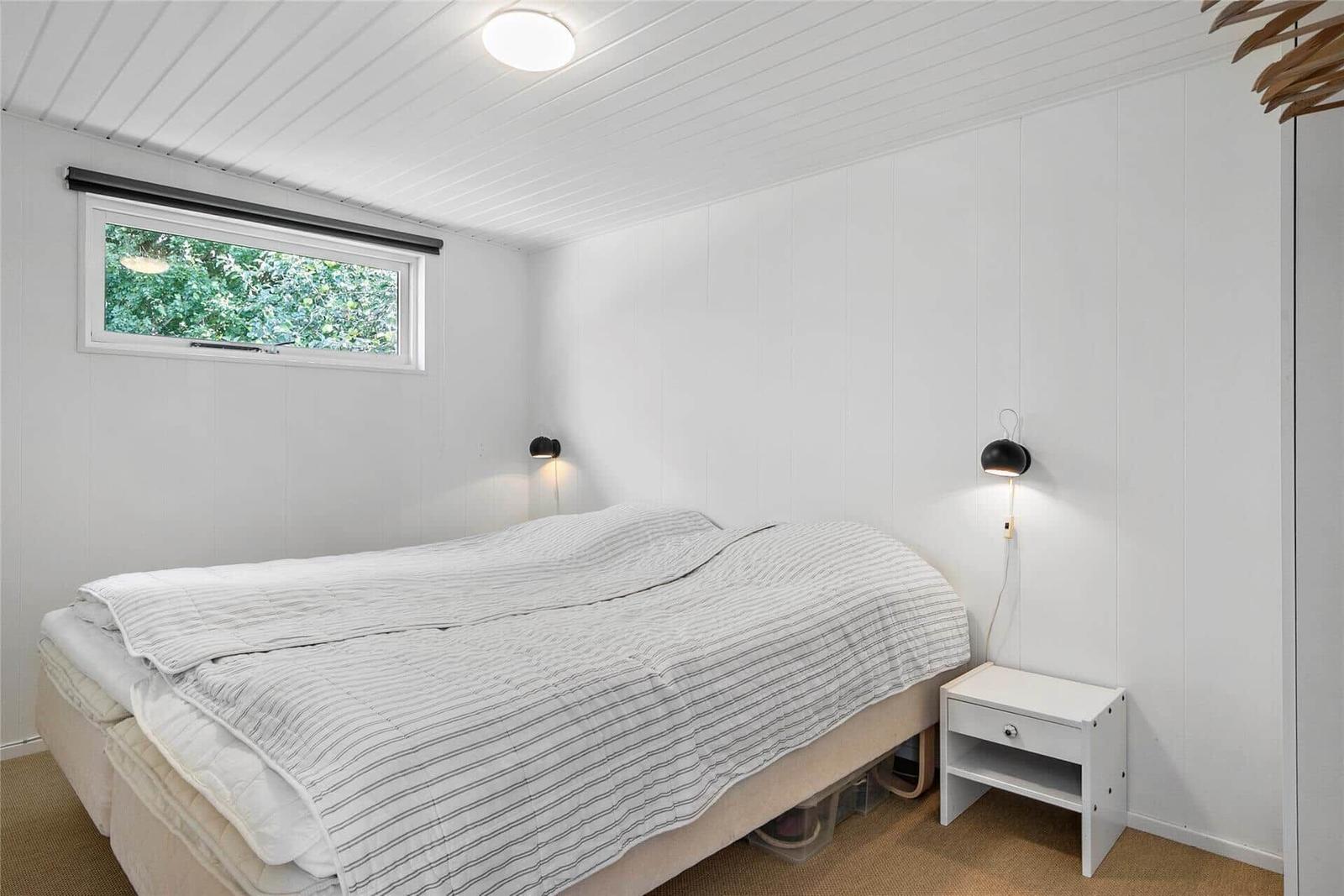 Schlafzimmer mit Bett, Nachttisch und Fenster. Wände und Decke sind weiß verkleidet.