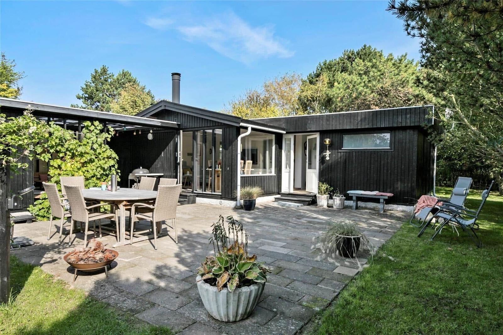 Schwarzes Holzhaus mit Terrasse, Gartenmöbeln und Grillplatz. Umgeben von Bäumen und Gras.