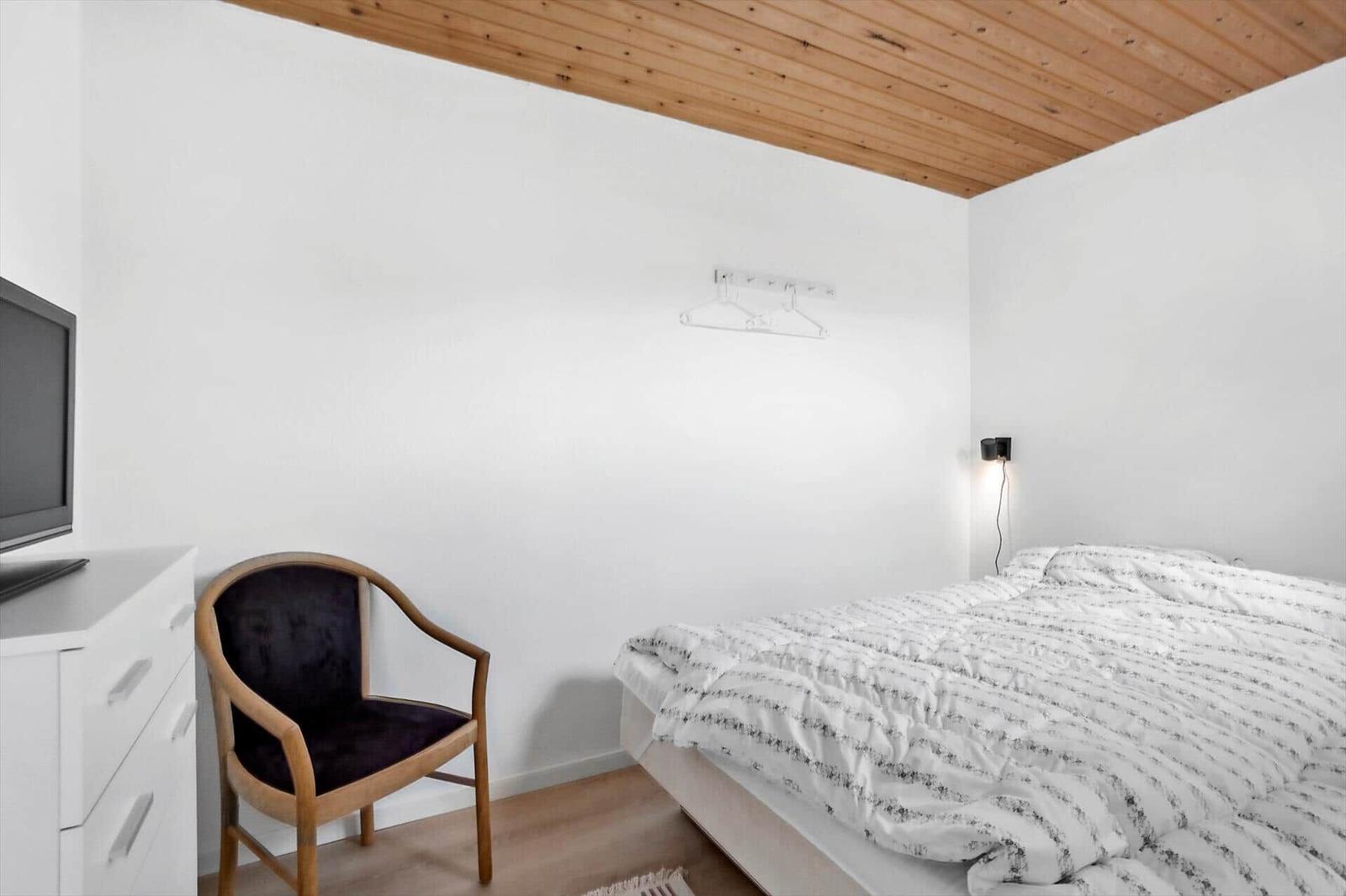 Schlafzimmer mit Bett, Stuhl und Fernseher an weißer Wand unter Holzdecke.