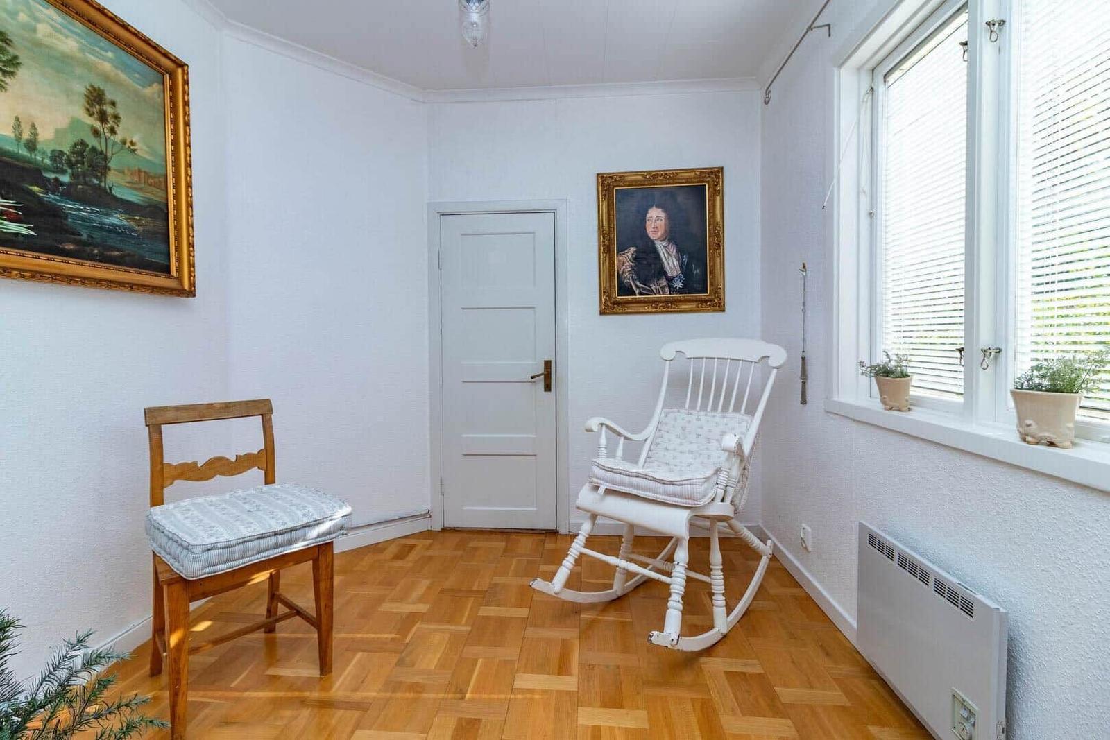 Raum mit Holzboden, weißer Schaukelstuhl und Holzstuhl, Wandgemälde und Fenster mit Jalousien.
