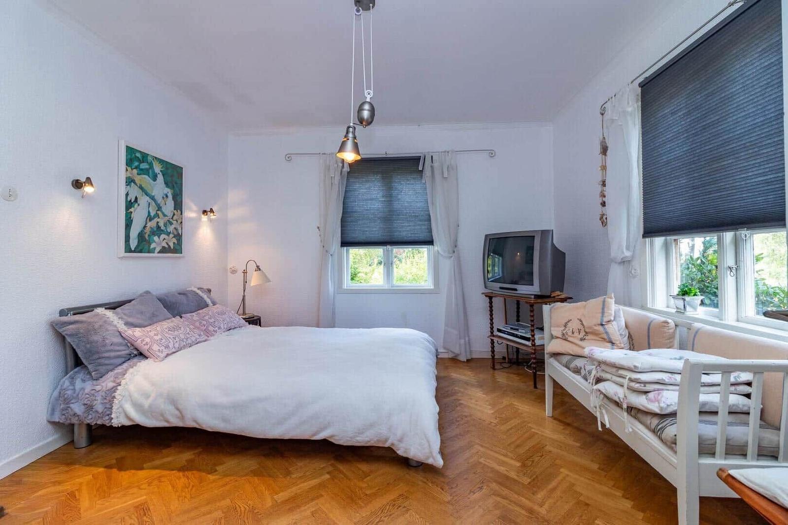 Schlafzimmer mit Bett, Sofa, TV und Holzboden. Fenster mit Jalousien und Wandbild.