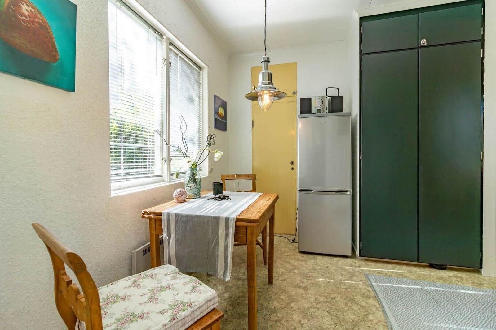 Essbereich mit Holztisch, Stühlen und Kühlschrank. Fenster mit Jalousien und grüner Schrankwand.