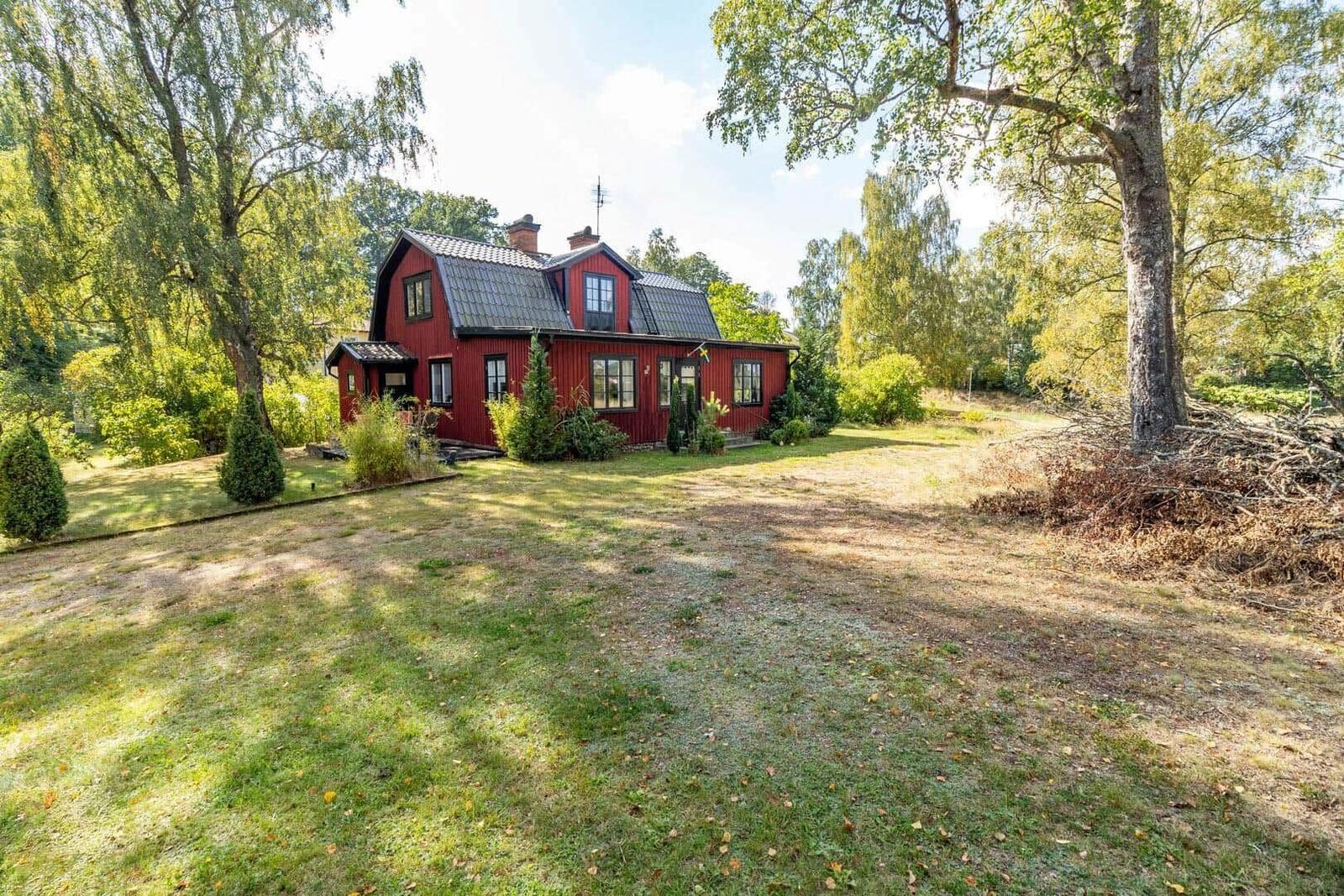 Rotes Haus mit dunklem Dach, umgeben von Bäumen und Grünfläche.