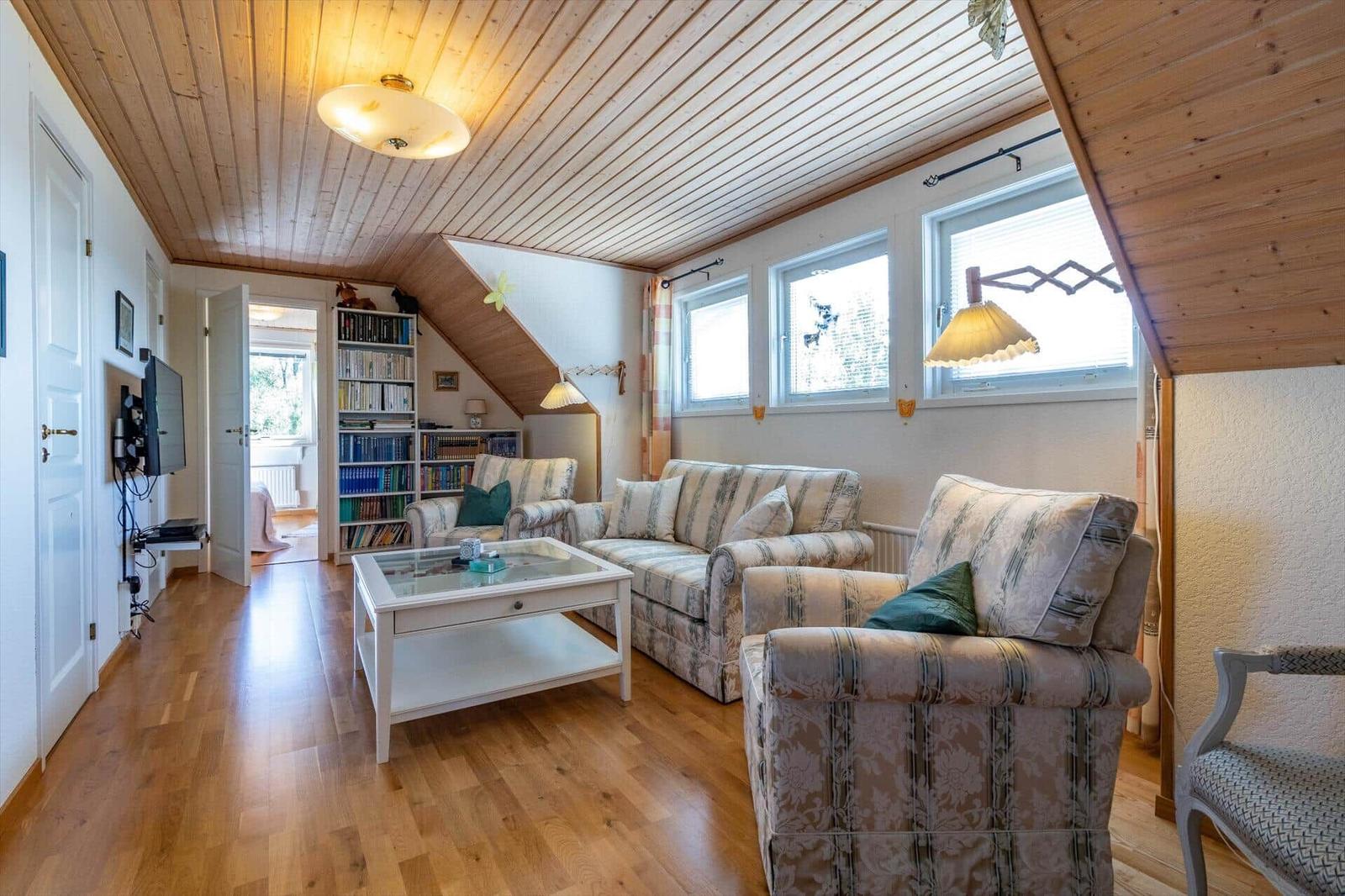 Wohnzimmer mit Holzdecke, Sofa, Sessel und Glas-Tisch. Bücherregal und Fenster sichtbar.