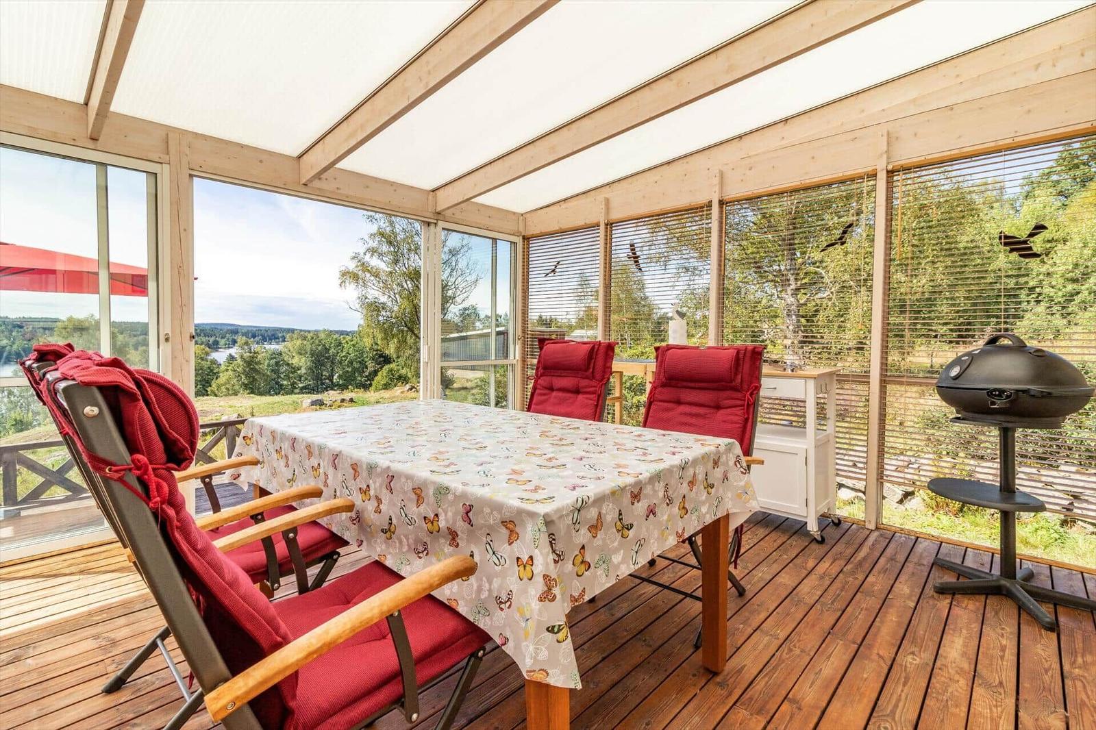 Terrasse mit Tisch, roten Stühlen und Grill. Blick auf Wald und See.