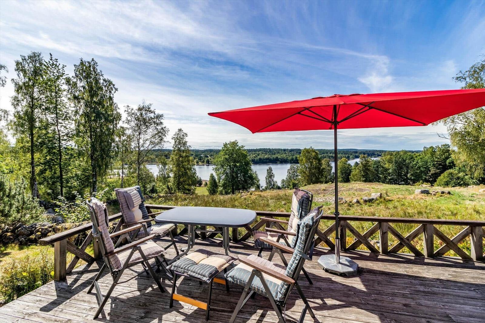 Terrasse mit Tisch, Stühlen und rotem Sonnenschirm. Blick auf See und Wald.