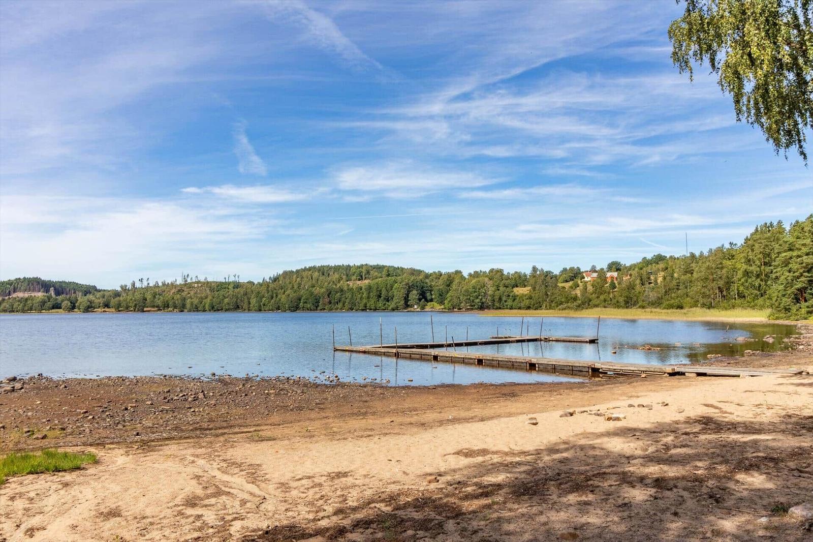 Seeufer mit Steg, Sandstrand und Wald im Hintergrund unter blauem Himmel.