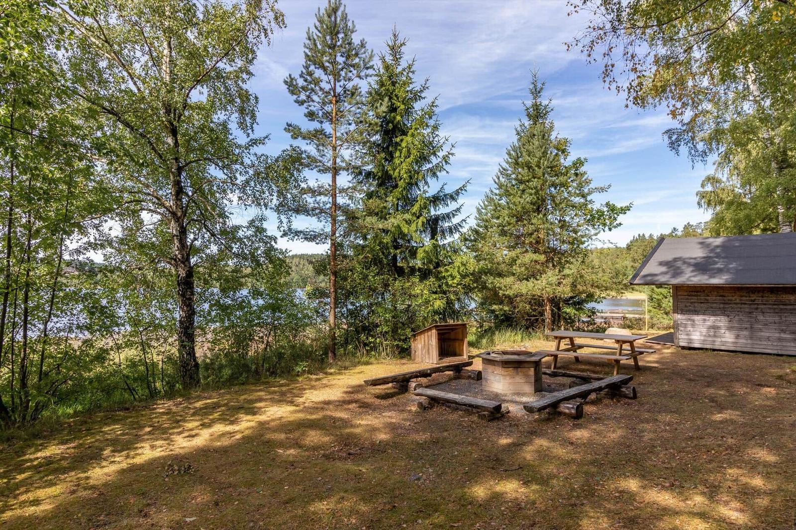 Hinter einem Holzgebäude steht ein runder Grillbereich mit Holzbänken und Tisch unter Bäumen.