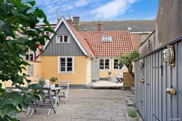 Hinterhof mit Tisch, Stühlen und Terrasse. Haus mit gelbem und grauem Holz. Dach mit roten Ziegeln.
