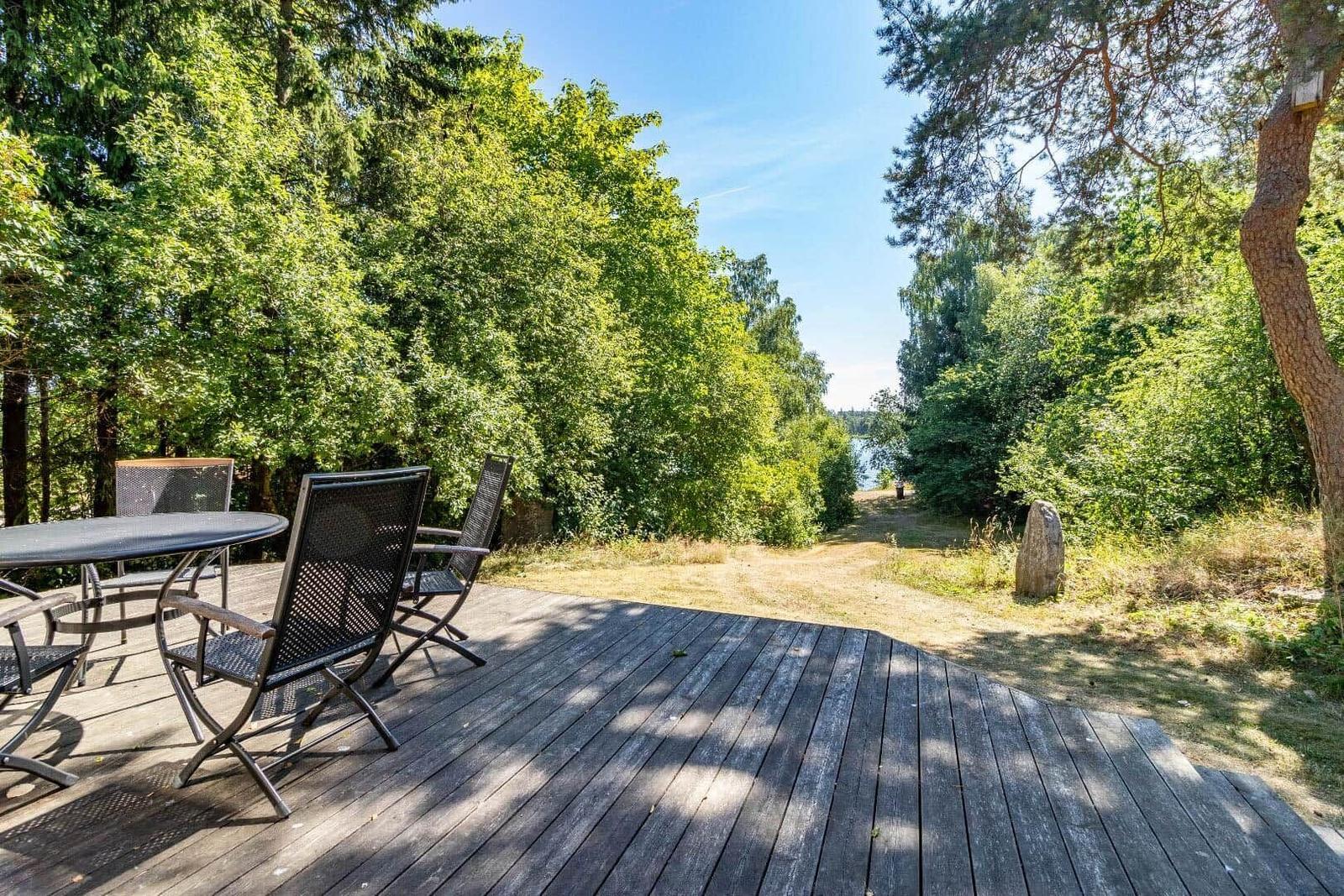 Holzterrasse mit Tisch und Stühlen, Blick durch Bäume zum Wasser.