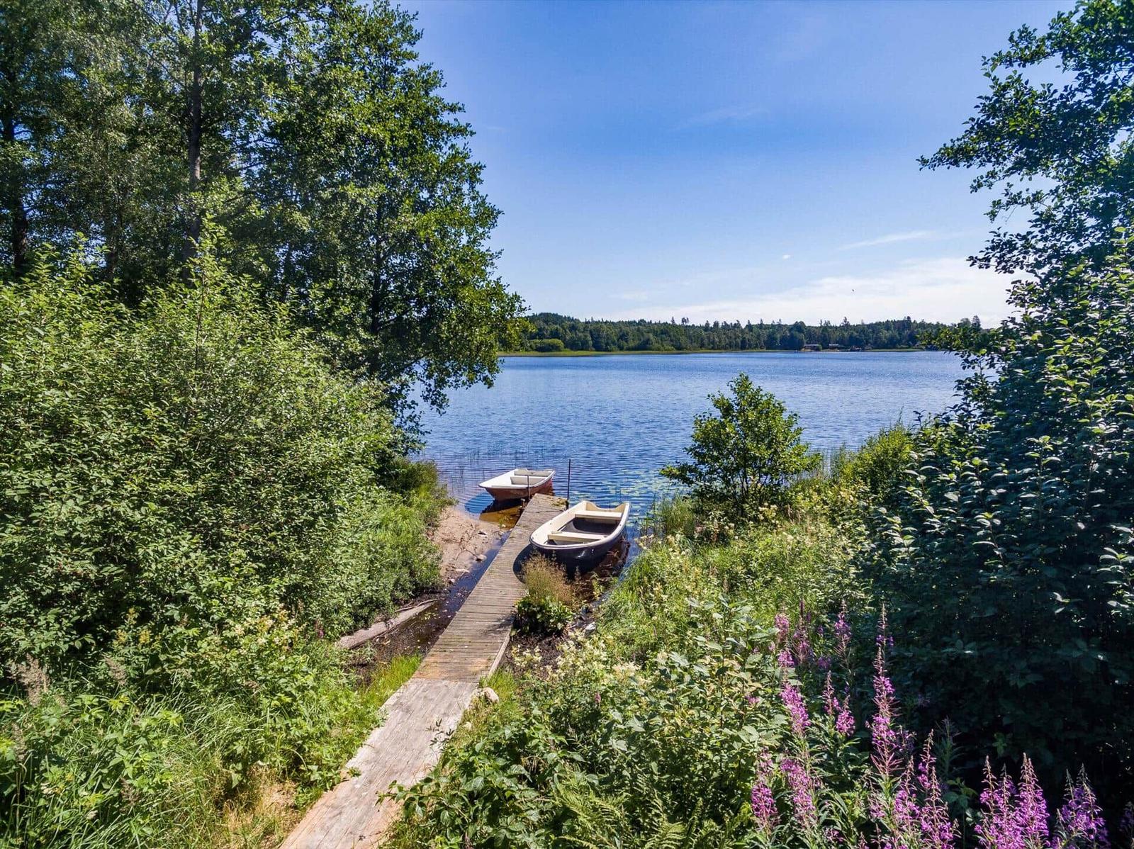 Hafen mit zwei Booten und Holzsteg am Seeufer. Umgeben von Grünflächen und Blumen.
