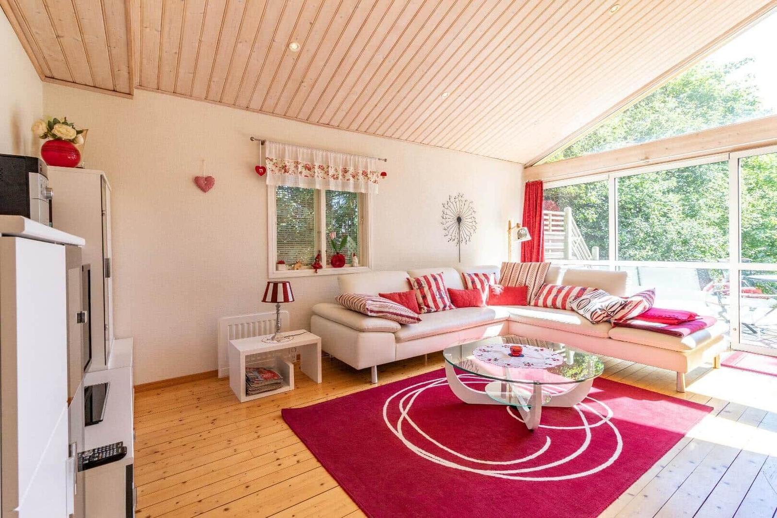 Wohnzimmer mit weißem Sofa, rotem Teppich und Glas-Tisch. Holzdecke und große Fenster.