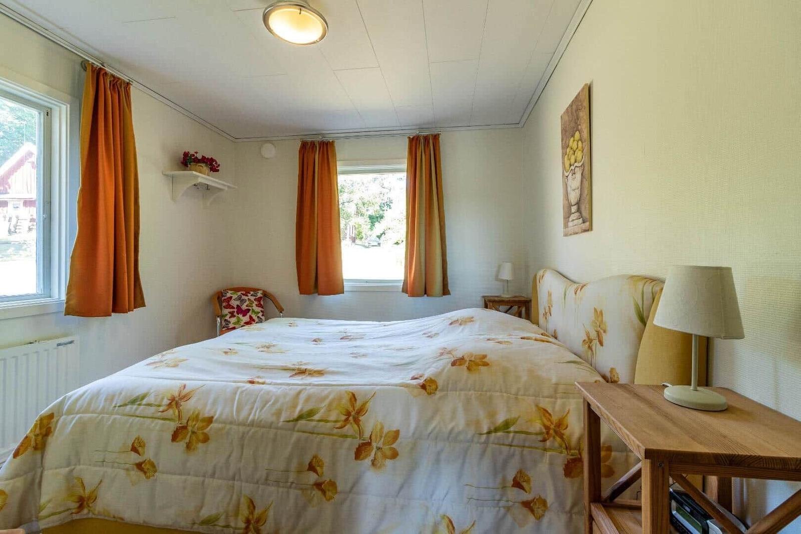 Schlafzimmer mit Bett, Nachttisch und Fenstern mit orangefarbenen Vorhängen.