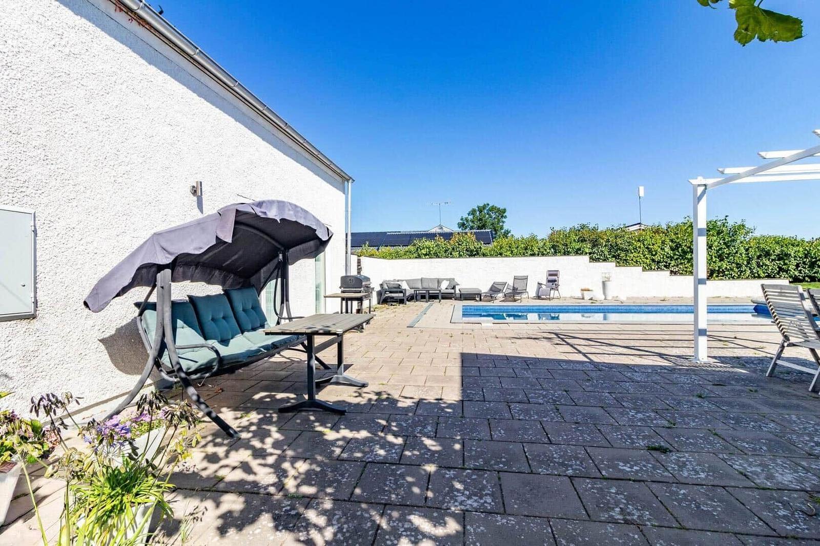 Terrasse mit Pool, Sitzgruppe und Grillplatz. Weiße Mauer und grüne Hecke im Hintergrund.