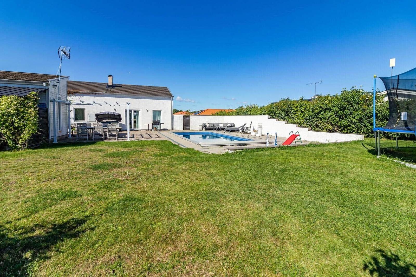 Haus mit Pool, Grillplatz und Trampolin auf grünem Grundstück unter blauem Himmel.