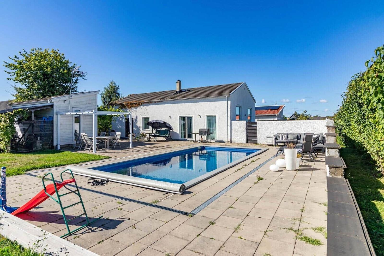 Haus mit Pool, Terrasse und Gartenmöbeln unter blauem Himmel.
