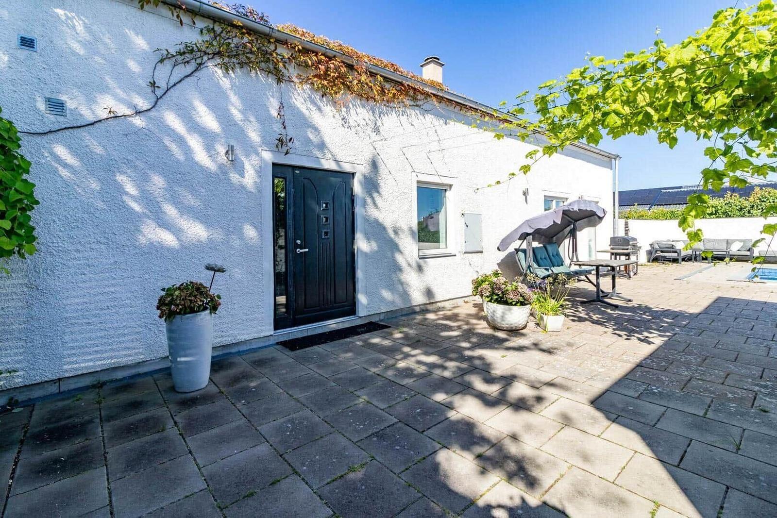 Haus mit Terrasse, Gartenmöbeln und Pool. Eingangstür und Pflanzen sind sichtbar.