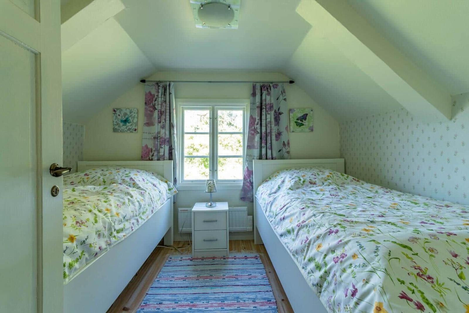 Zwei Betten mit floraler Bettwäsche stehen unter einem Dachbodenfenster mit Vorhängen.